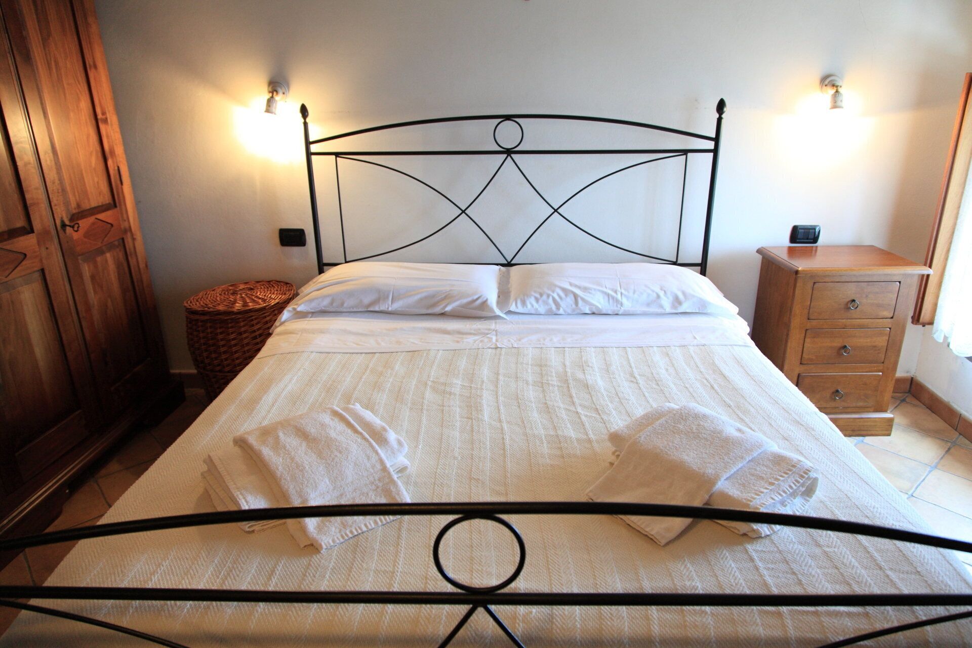A neatly made bed with white linens and towels, a wooden wardrobe, a wicker basket, and a wooden nightstand with drawers.