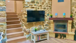 A stone-walled room with a fireplace, a flat-screen TV mounted on the wall, a small table with books, and a staircase leading to another room. - LA CASA DEL RÍO. Ideal for families with children and couples seeking tranquility (Pujayo)