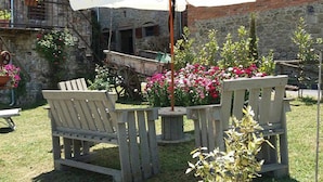 A rustic outdoor seating area with wooden chairs and a white umbrella, surrounded by flowering plants and a stone building with a sign reading "Antica Fabbrica del Tabacco".