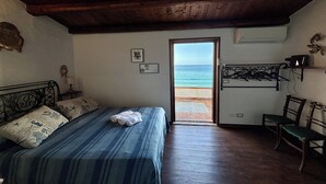 Quarto com cama, poltrona, porta para varanda e vista para o mar, e uma mesinha com luminária.