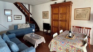 Uma sala de estar aconchegante com um sofá azul, uma mesa de jantar de madeira com toalha de mesa estampada, e uma escada que leva a um andar superior.
