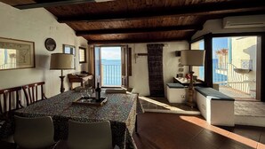 Uma sala de jantar com mesa posta para quatro pessoas, teto de madeira e vista para o mar por uma porta aberta.