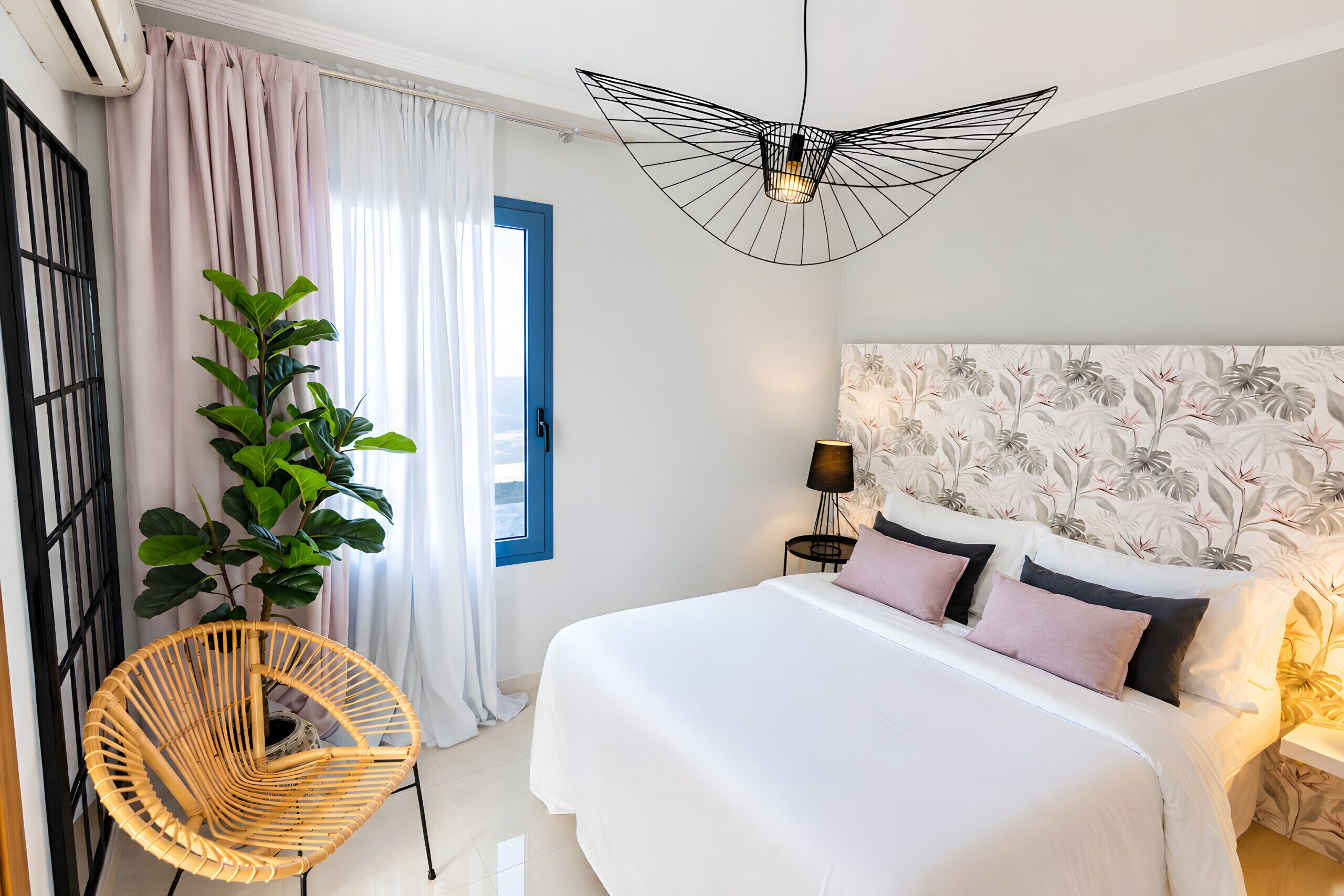 Une chambre à coucher avec un lit, un luminaire suspendu, une plante en pot, une chaise et une fenêtre avec des rideaux.