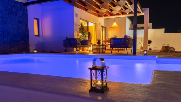 An outdoor swimming pool area with a stone wall, a wooden pergola, and a seating area with chairs and a table. - Villa Arai   (Playa Blanca)