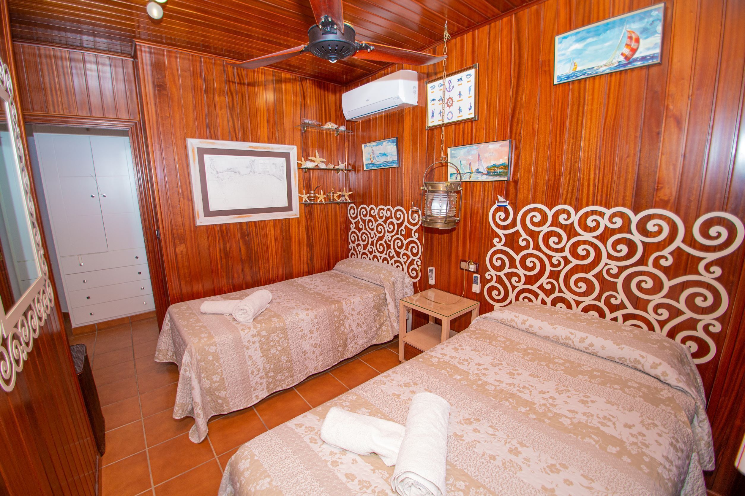 A hotel room with wooden paneling, two beds with matching headboards, a ceiling fan, an air conditioning unit, and a small table with a lantern.