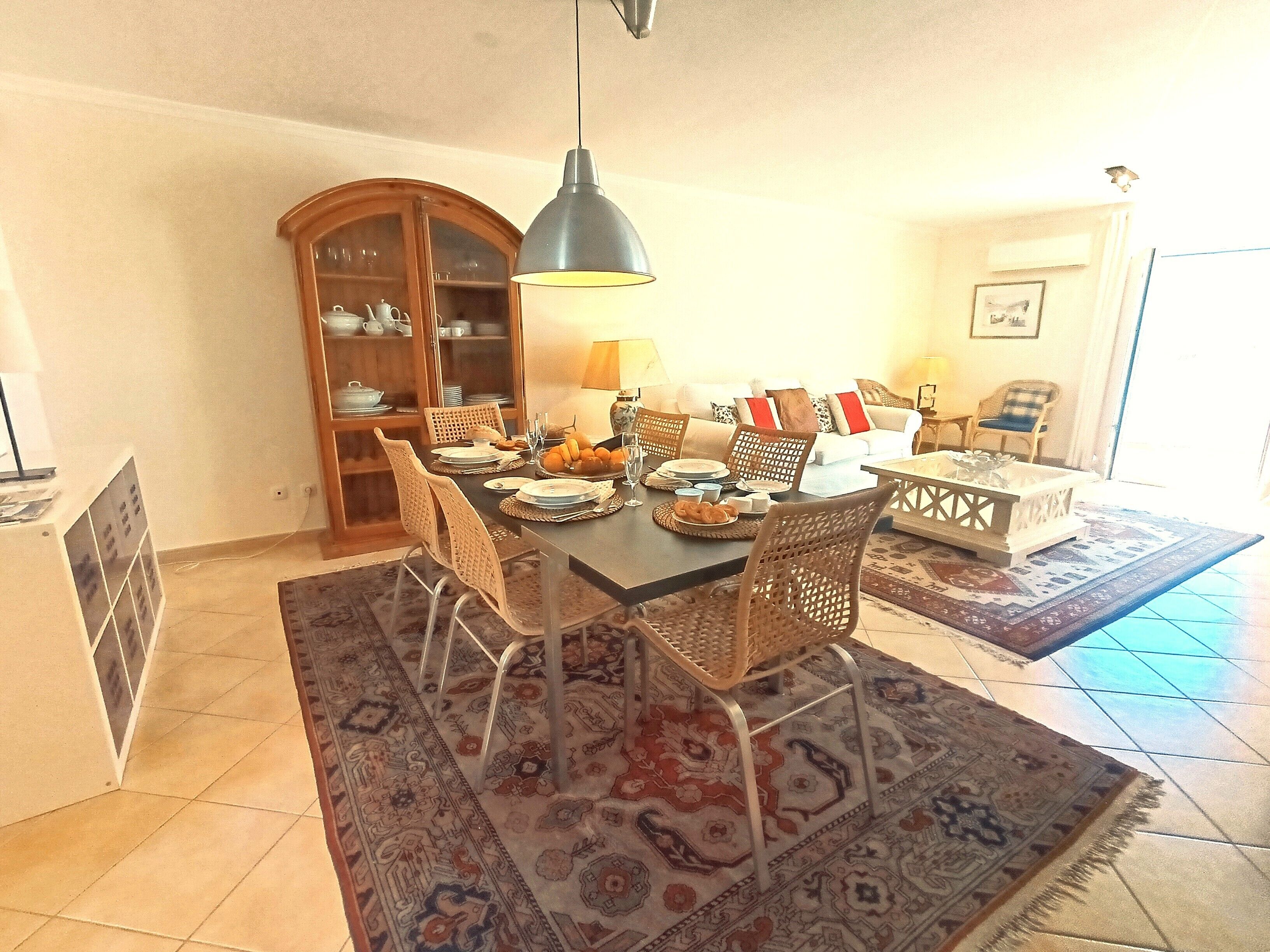 A dining area with a table set for four, surrounded by wicker chairs, a patterned rug, and a cabinet with dishes.