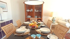 Een eettafel gedekt met borden, glazen en eten, omringd door rieten stoelen en een kast met servies.