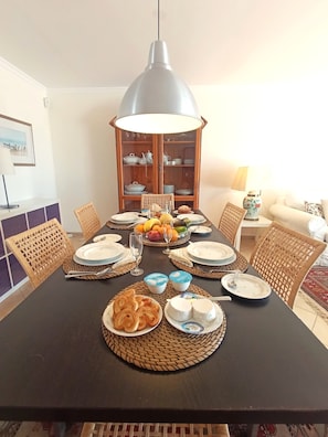 A dining table set with plates, glasses, and food, surrounded by wicker chairs and a cabinet with dishes.