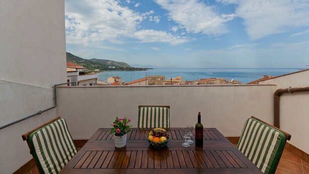 A balcony with a wooden table, chairs, a vase of flowers, a bottle of wine, and a view of the sea and buildings. - Terrazza del Centro (Cefalù)