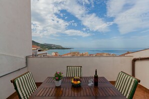 A balcony with a wooden table, chairs, a vase of flowers, a bottle of wine, and a view of the sea and buildings.