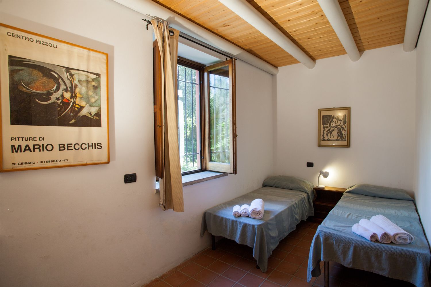 Una stanza con due letti, una finestra con tende, un quadro appeso al muro e un piccolo tavolo con asciugamani.