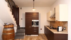 A compact kitchen with dark wooden cabinets, a built-in oven, and a stainless steel range hood. A wooden barrel and a basket are placed near the stairs. - Wine House (Cefalù)