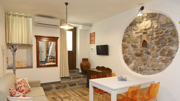 A living room with a stone wall, a white table, orange chairs, a sofa, a TV, and a wooden cabinet. - Wine House (Cefalù)