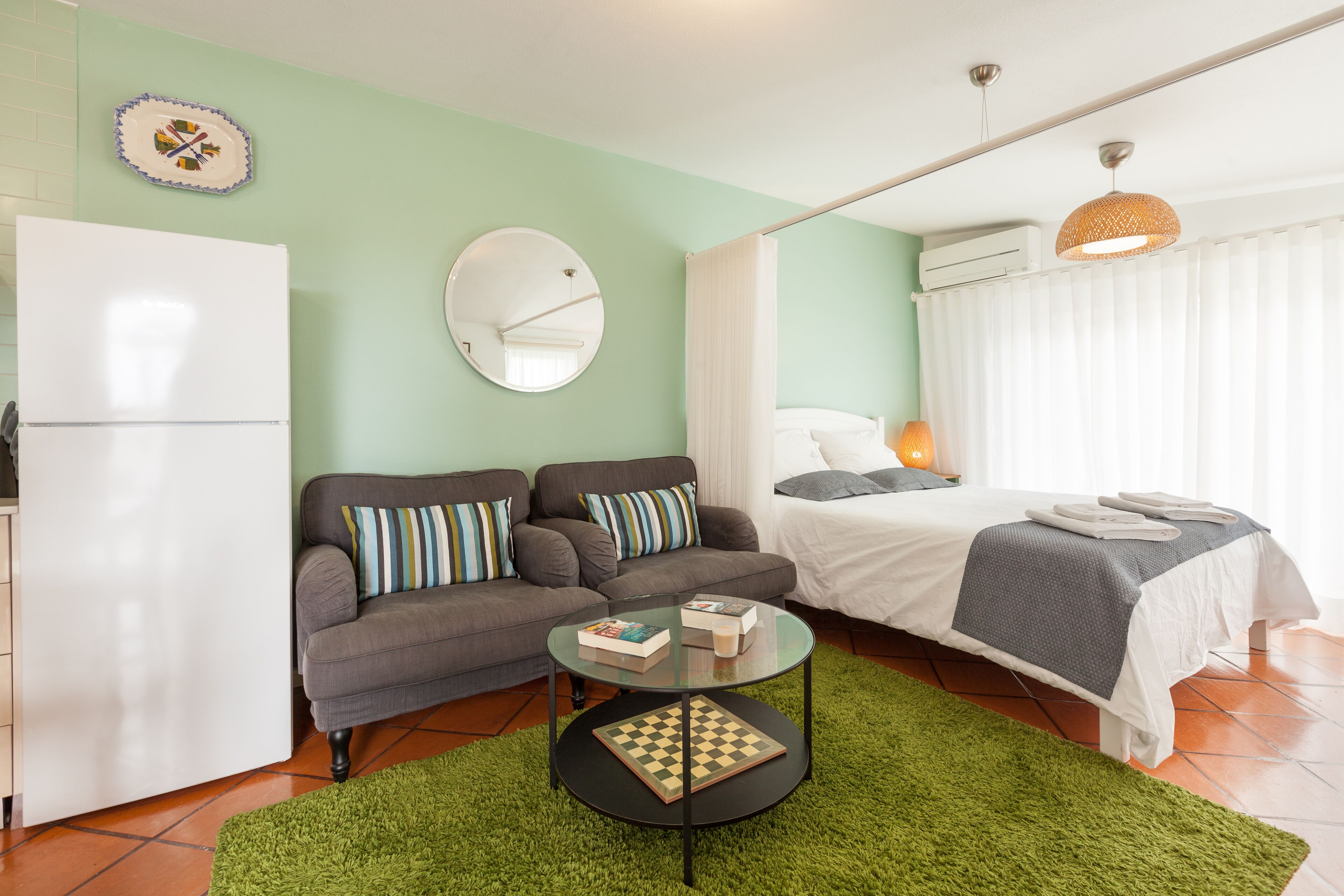 Een knus woongedeelte met een bank, een ronde tafel met boeken, een bed met een witte dekbedovertrek en een groen vloerkleed.