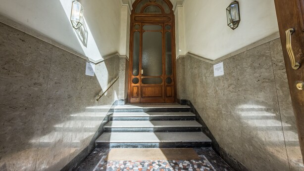 Eine Marmortreppe führt zu einer Holztür mit Glasfenster, begleitet von Wänden mit strukturierter Oberfläche und Wandleuchten.