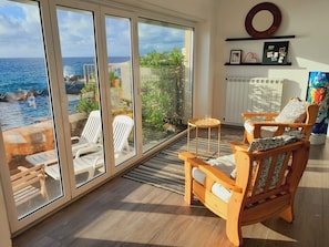 Une chambre avec vue sur l’océan, comprenant un coin salon avec des fauteuils et une petite table, un radiateur et une étagère ornée d’objets décoratifs.