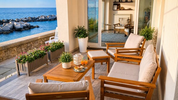 A sunlit outdoor seating area with wooden furniture, potted plants, and a view of the sea.