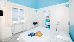 Une chambre avec un grand lit, une valise et un chapeau posés sur le lit. Il y a une fenêtre avec des rideaux, un radiateur et une vue sur la mer.