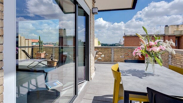 Een modern terras buiten, met glazen wand, een tafel met stoelen en een bloemenvaas.