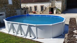 Une piscine hors sol dotée d’une structure blanche, entourée d’une terrasse en béton, située devant un bâtiment à la façade en pierre et au toit recouvert de tuiles.
