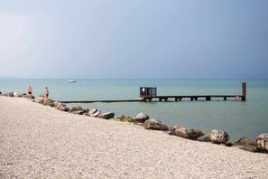 A pebbled beach with a wooden pier extending into calm waters, a small wooden structure at the end, and a clear sky. - Residence -2 Saint Benedict of Lugana (Peschiera Del Garda)