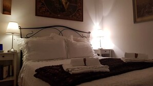 En snyggt bäddad säng med vita sängkläder och en mörk pläd, två sänglampor, ett litet nattduksbord med en lampa och en inramad tavla ovanför sängen.
