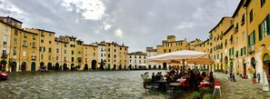 A cobblestone square with outdoor seating, surrounded by yellow buildings with green shutters.