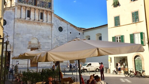 Un café al aire libre con mesas y sillas, una fuente y un edificio histórico con un mosaico al fondo.