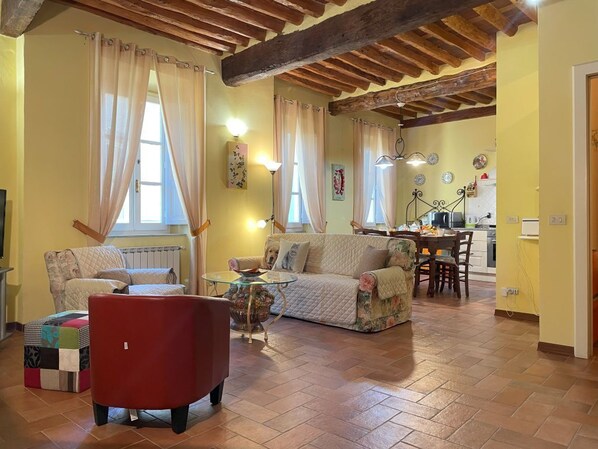 A cozy living room with a wooden ceiling, a red armchair, a floral sofa, and a dining area with a table and chairs.