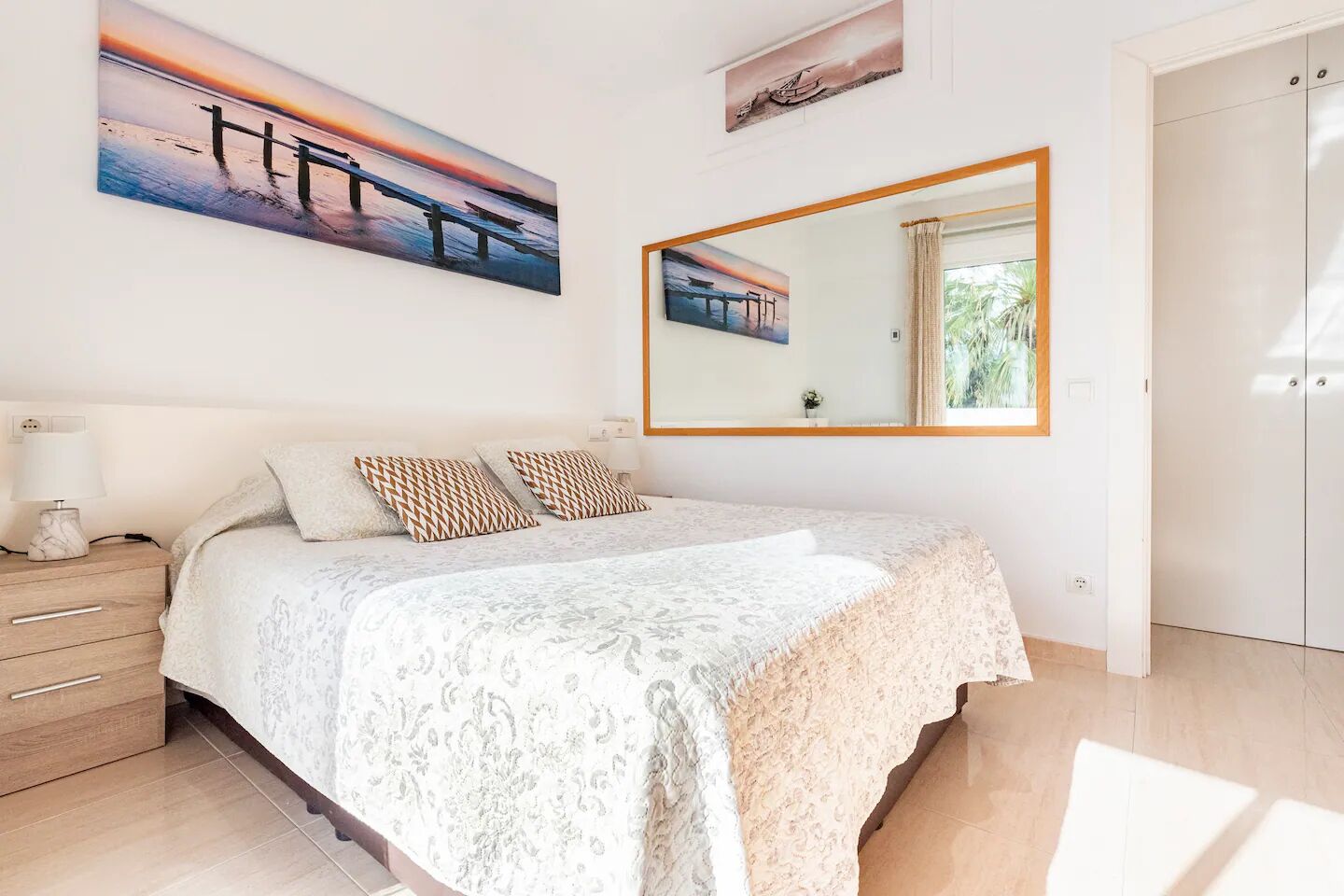 A bedroom with a bed, a nightstand, a mirror, and artwork on the wall.