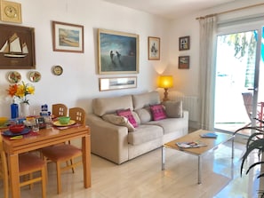 A living room with a sofa, coffee table, dining table, and chairs. There are framed pictures on the walls, a lamp, and a window with curtains.