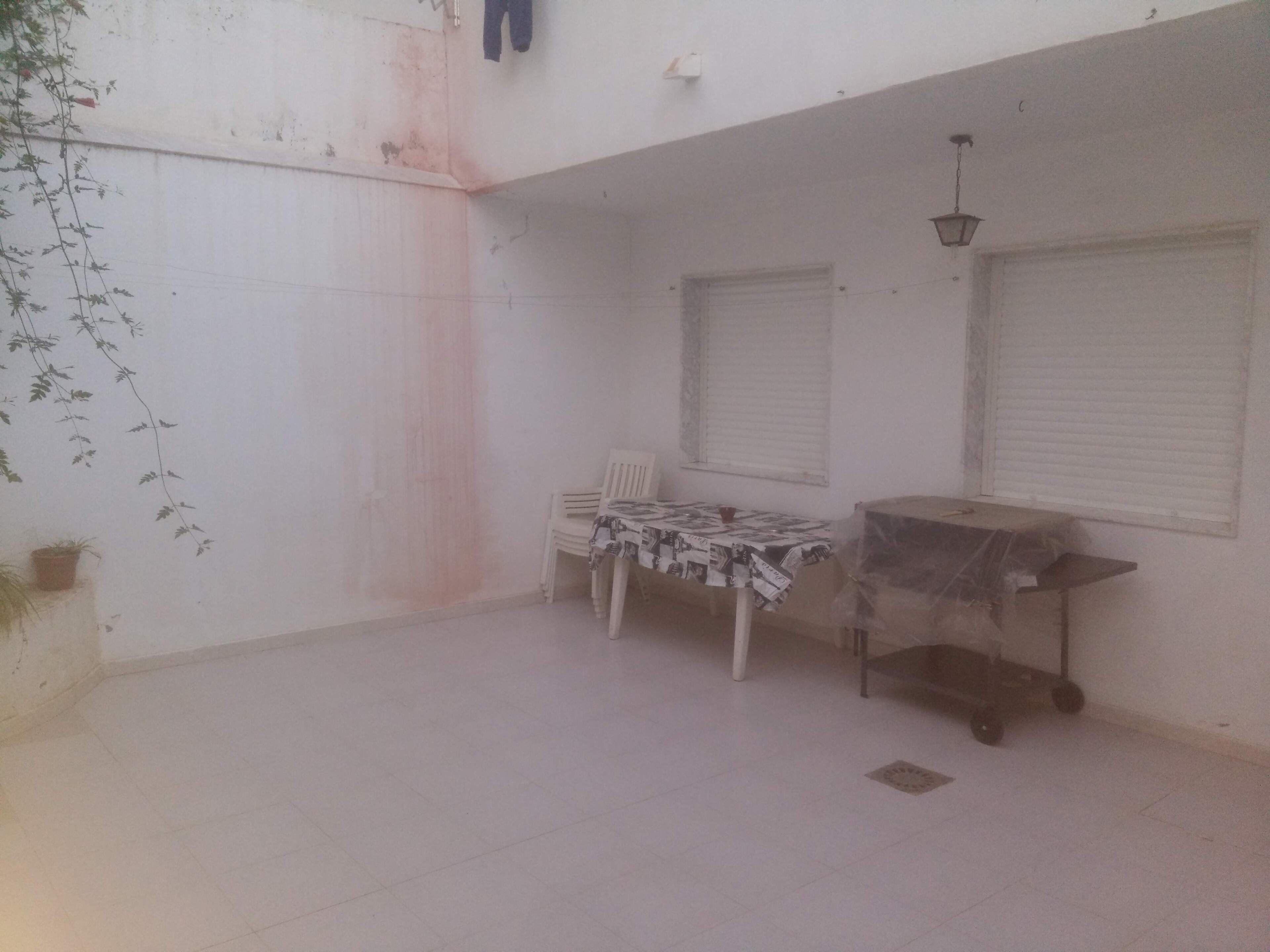 A small, enclosed patio area with a tiled floor, a table with a patterned tablecloth, a chair, and a covered cart.