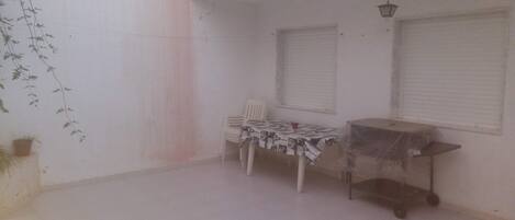 A small, enclosed patio area with a tiled floor, a table with a patterned tablecloth, a chair, and a covered cart.