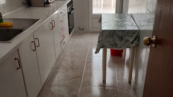 Et køkken med hvide skabe, en vask og et bord med dug med bladmotiv.