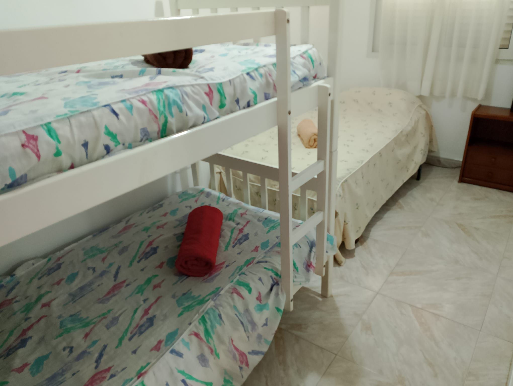 Een stapelbed met een gestreept matras, een klein houten nachtkastje en een raam met jaloezieën.