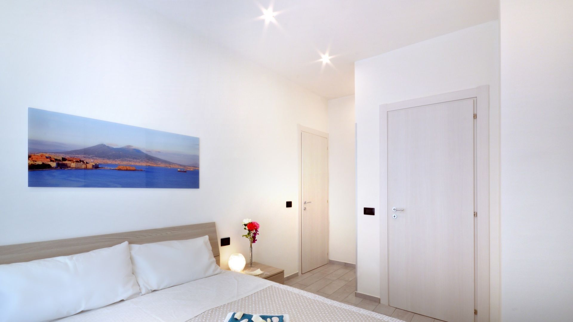 Un dormitorio con cama, mesita de noche con lámpara, una puerta y un cuadro colgado en la pared de un paisaje costero.