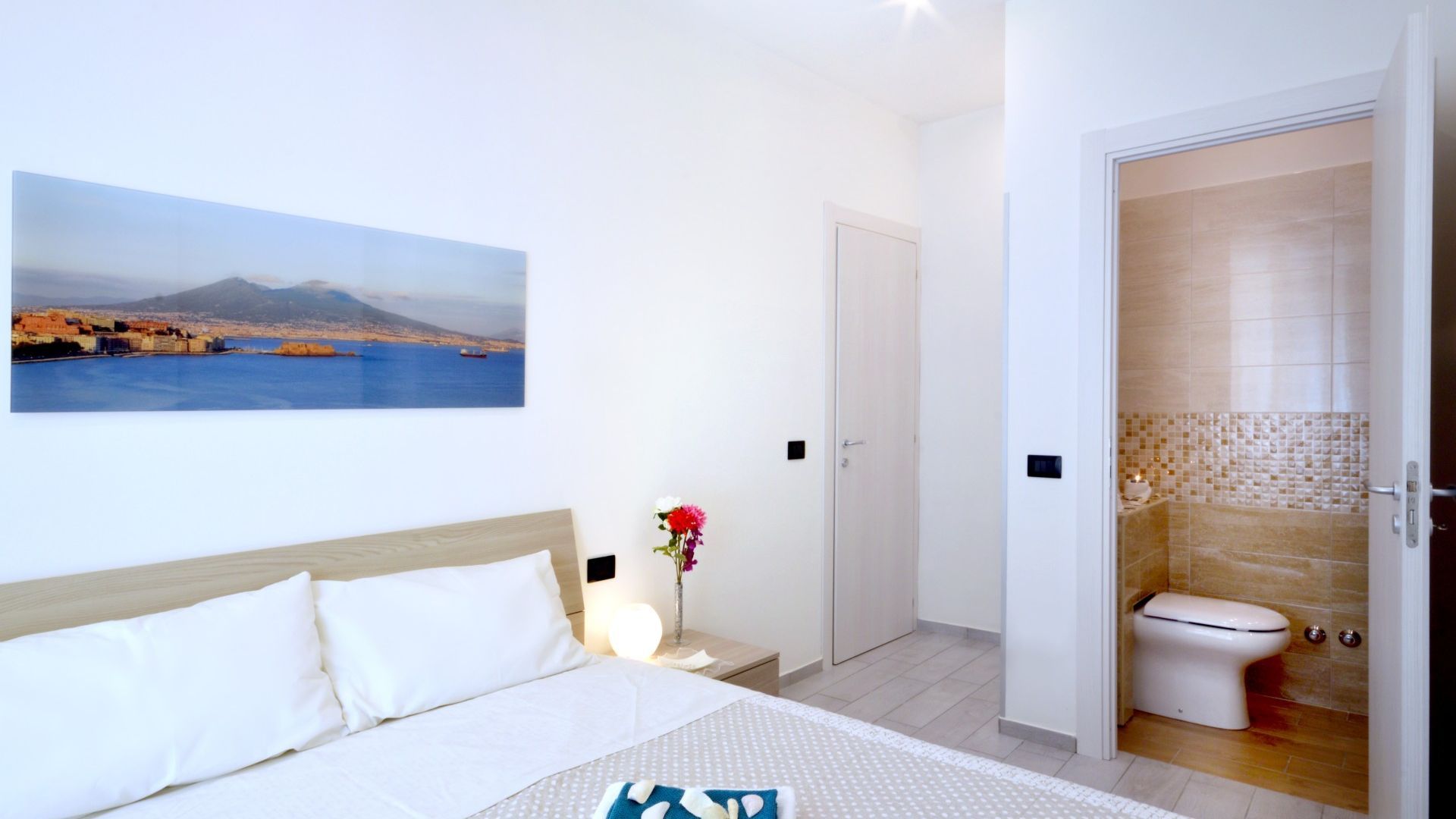 Une chambre d’hôtel avec un lit, un tableau représentant un paysage côtier, et une salle de bain avec des toilettes et des murs carrelés.