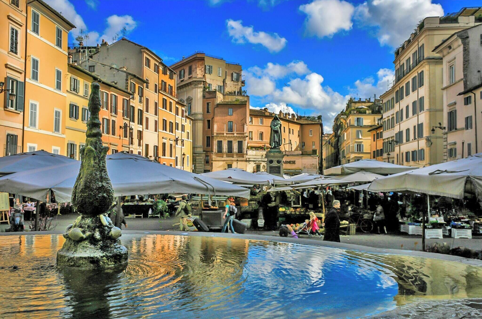 A public square with a fountain, market stalls, and historic buildings.