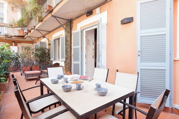 Un área de comedor al aire libre con una mesa lista para cuatro personas, rodeada de sillas, un porche cubierto con plantas y una barandilla.
