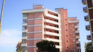 Un immeuble résidentiel à plusieurs étages, doté de balcons, avec un ciel dégagé et un arbre au premier plan.