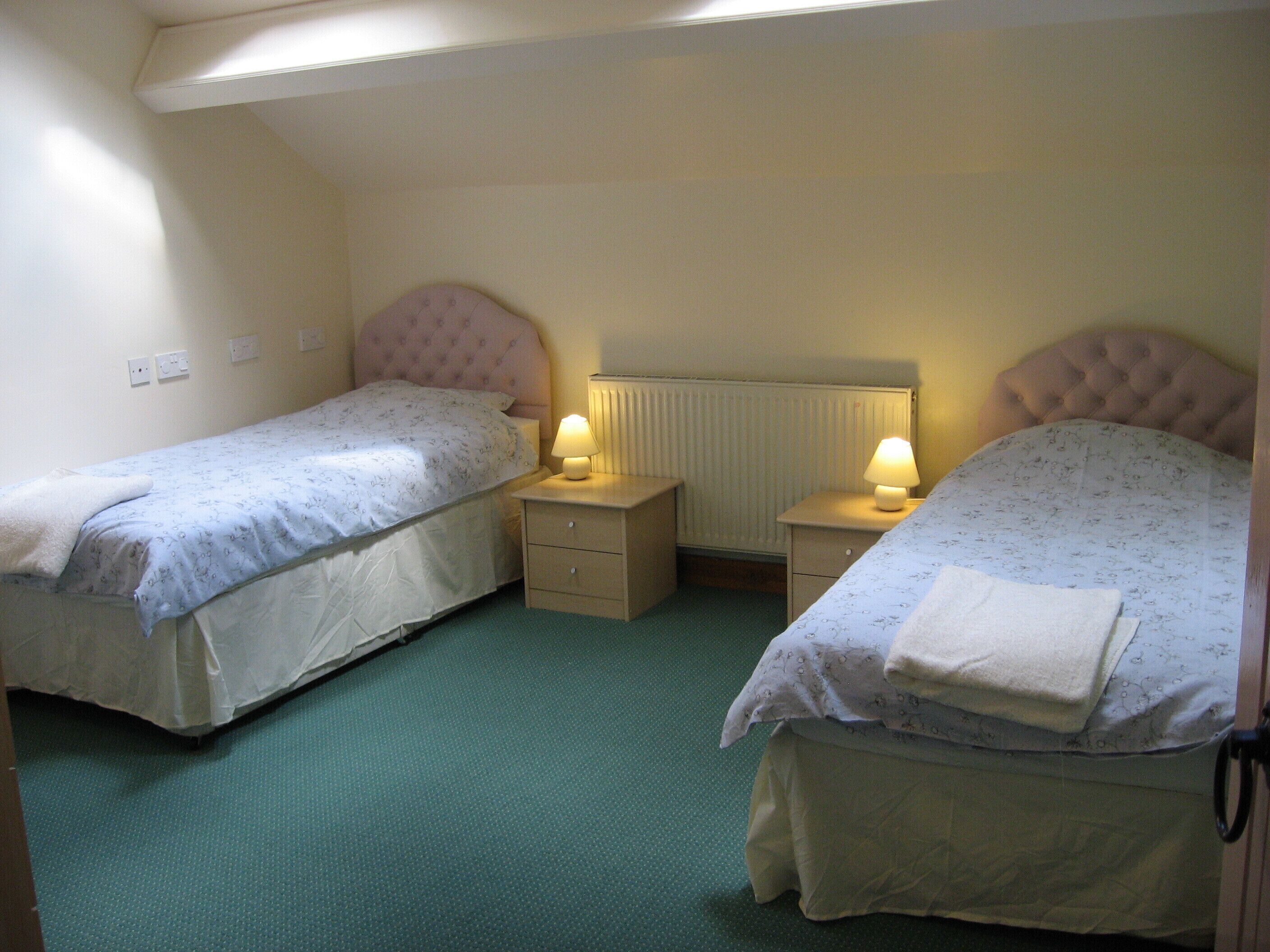 A small, cozy room with two single beds, each with a headboard and bedside tables with lamps. The room has a simple layout with a radiator above the beds and a green carpet.