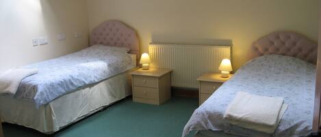 A small, cozy room with two single beds, each with a headboard and bedside tables with lamps. The room has a simple layout with a radiator above the beds and a green carpet.