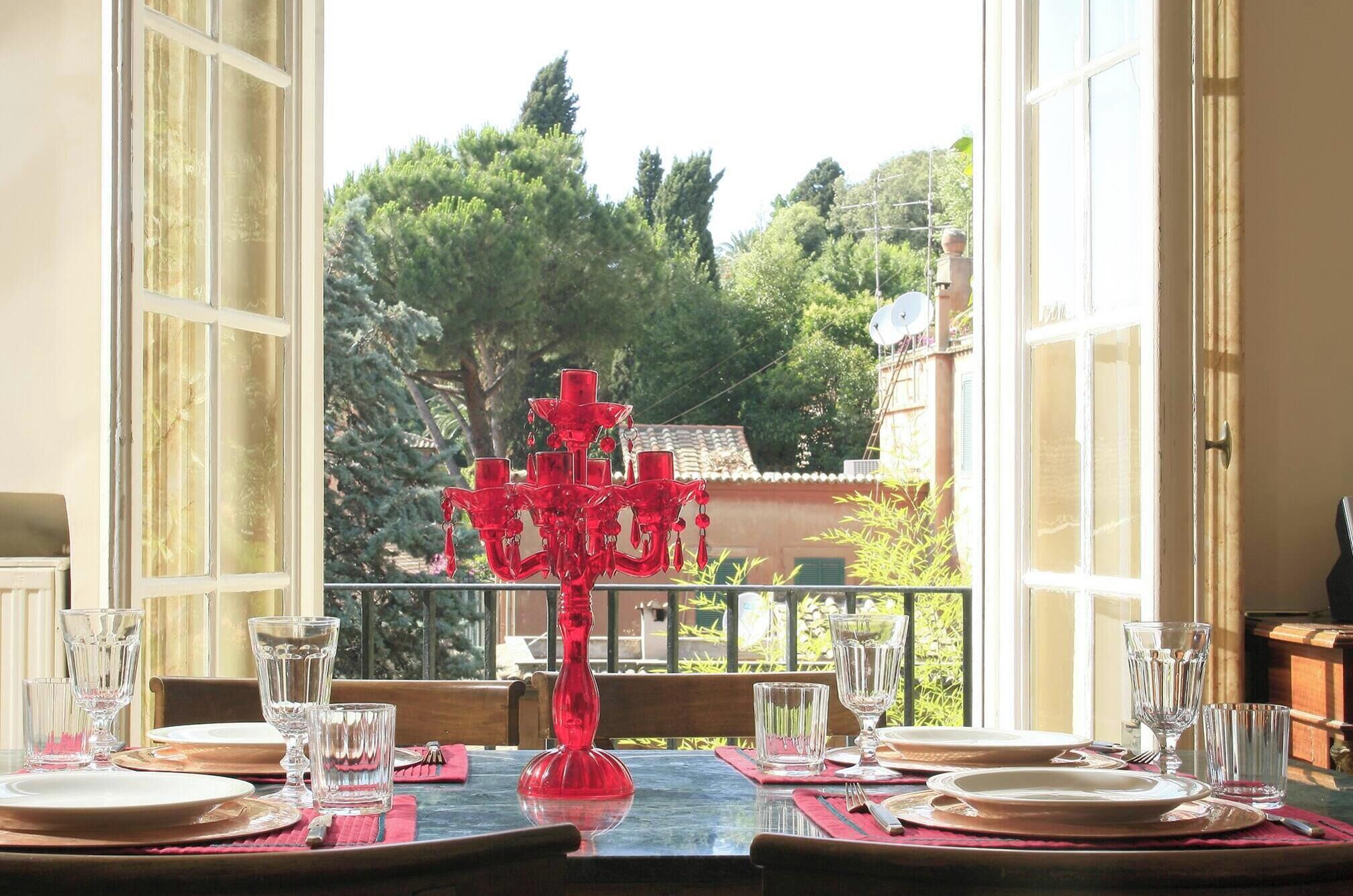 Mesa de jantar posta para quatro pessoas, com copos de vidro transparente e um centro de mesa de vidro vermelho, com vista para um jardim e edifícios.