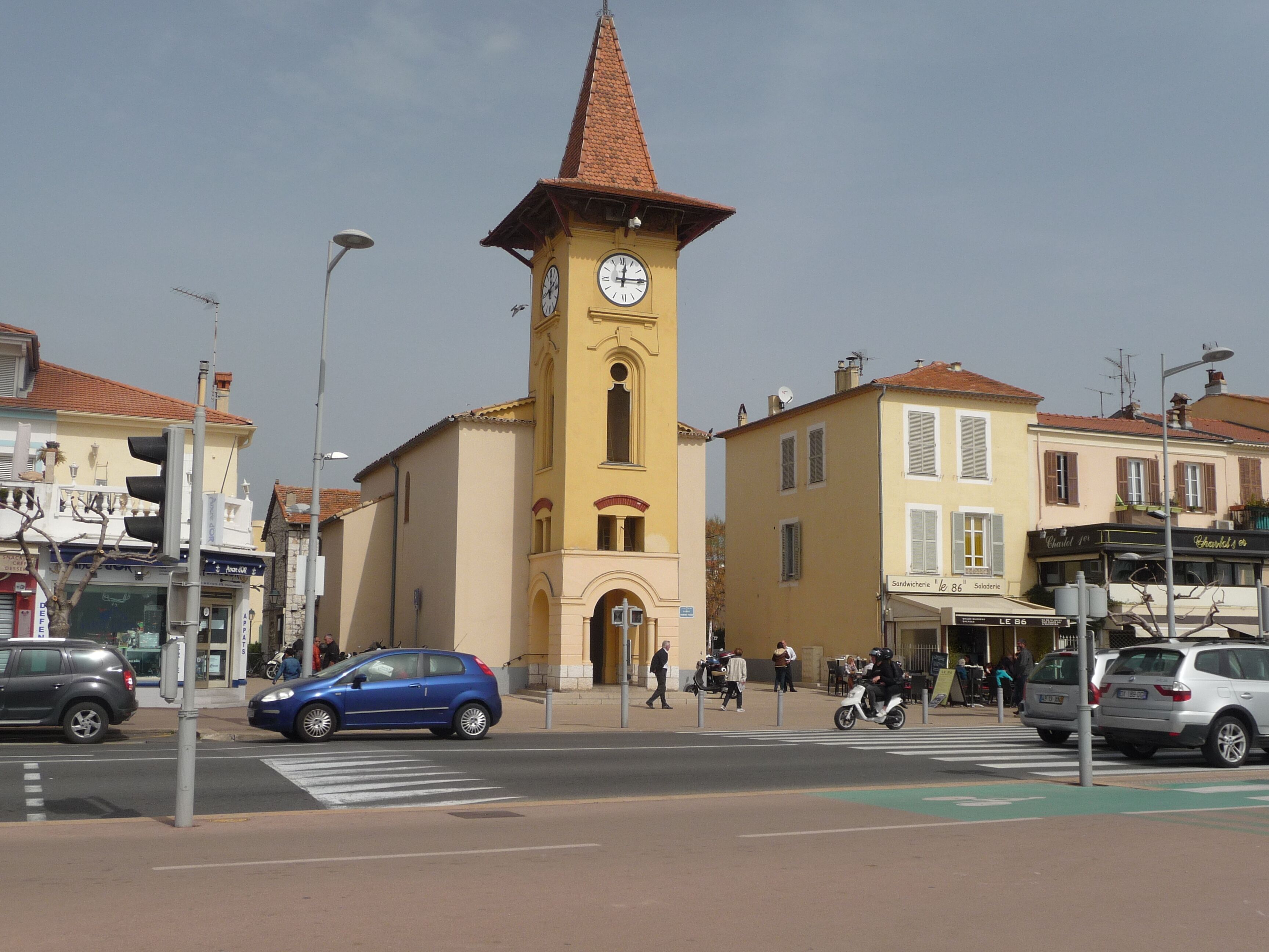 Un edificio amarillo con una torre de reloj, un paso de peatones y una calle con autos y peatones.