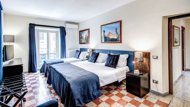 A hotel room with a checkered floor, a large bed with blue bedding, a desk with a lamp, and two paintings on the wall. - Times Rome Colosseum, wide in wonderful location front Coliseum (Rome)