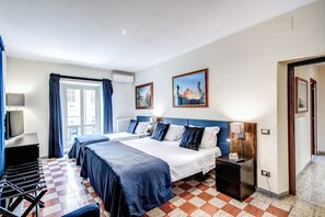Une chambre d’hôtel avec un sol à carreaux, un grand lit avec une literie bleue, un bureau avec une lampe et deux tableaux accrochés au mur.