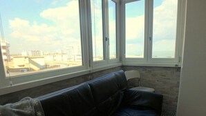 A room with a black leather sofa, a tiled wall, and a large window offering a view of buildings and a clear sky. - Sea view, bright apartment in the center of Lido di Pomposa (Comacchio)