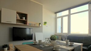 A modern living room with a flat-screen TV, a bookshelf, a dining table with cups and a pitcher, and a large window with blinds. - Sea view, bright apartment in the center of Lido di Pomposa (Comacchio)