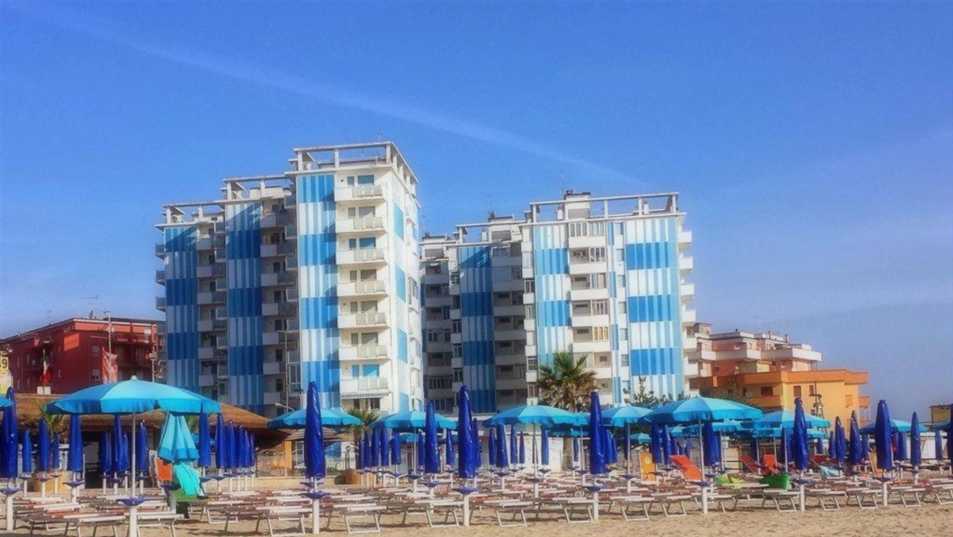 Un hotel frente a la playa con varios edificios de rayas azules, muchas sombrillas y sillas de playa.