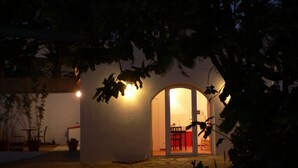 Un edificio con un arco de entrada que da a una habitación con una silla roja y una mesa. Hay plantas en macetas afuera y una lámpara fijada a la pared sobre el arco.
